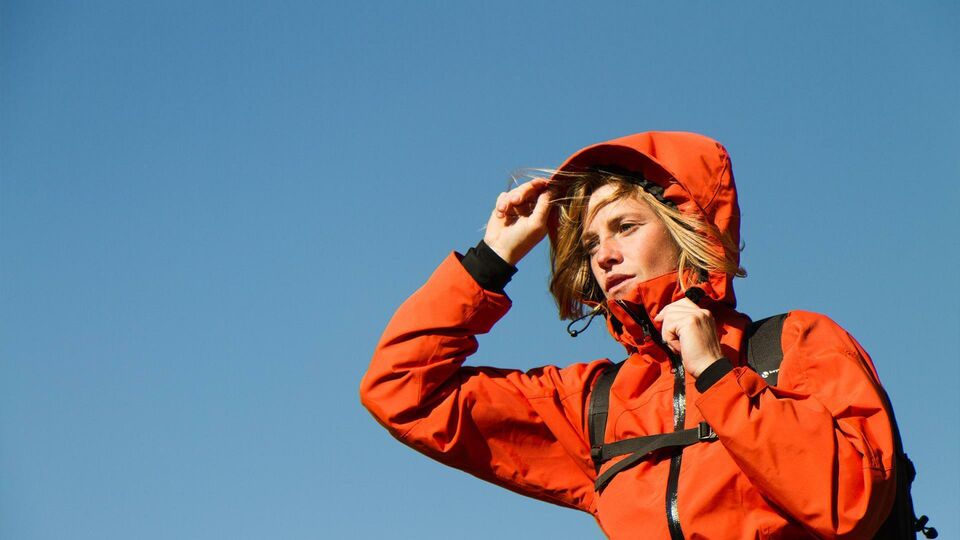 A young woman in red hiking jacket raises her hood, no other details can be seen except a clear blue sky.